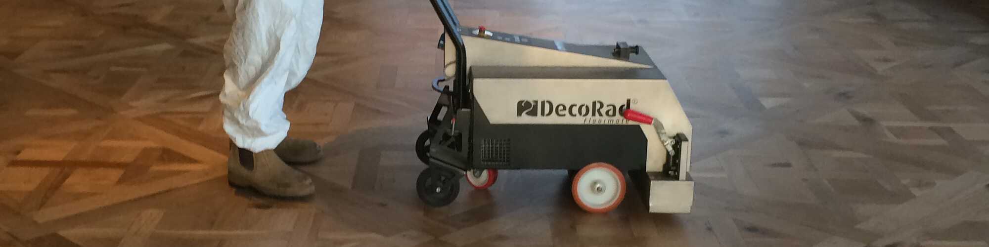 A person in white coveralls pushes a DecoRad machine on wooden flooring.