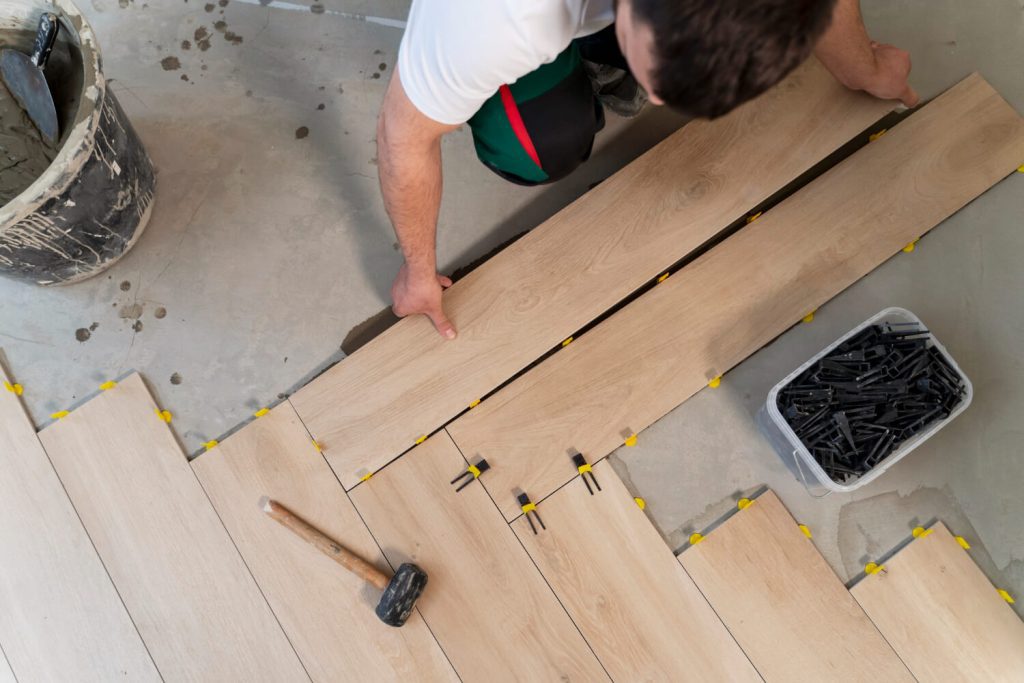 A person lays wooden floor planks using spacers and a rubber mallet on the floor.